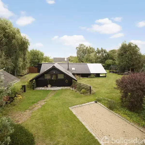 Et sort træhus står i en grøn have med sandareal og buske. To mindre bygninger ses bagved. Himlen er delvist skyet.