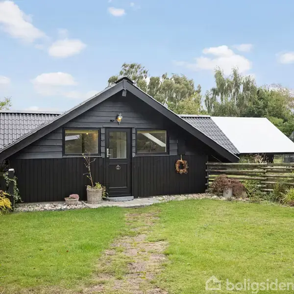 Et sort træhus med store vinduer står i en grøn have med en lille sti og blomsterkrukke ved hoveddøren. I baggrunden ses træer og buske.
