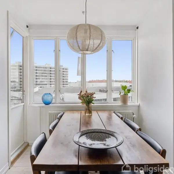 Træbord med tallerken og vase med blomster står centralt. Omgivet af stole, foran store vinduer med byudsigt. En loftlampe hænger over bordet. En blå vase og plante er i vindueskarmen.
