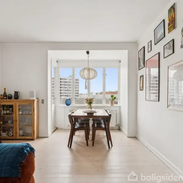Spisebord med stole står i en lys spisestue, dekoreret med kunst på væggen. Et vitrineskab til venstre med et vinreol. Store vinduer viser byudsigt.
