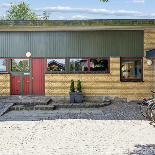 En gul murstensbygning med røde og grønne dørpaneler og store vinduer. Flere cykler står parkeret foran på det stenbelagte fortov, omgivet af træer og buske.
