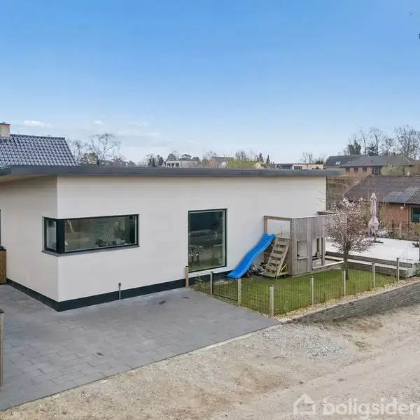 En moderne, hvid bygning med fladt tag har en tilknyttet terrasse og en blå rutsjebane. Bygningen ligger i et villakvarter med omkringliggende huse og en grusvej.