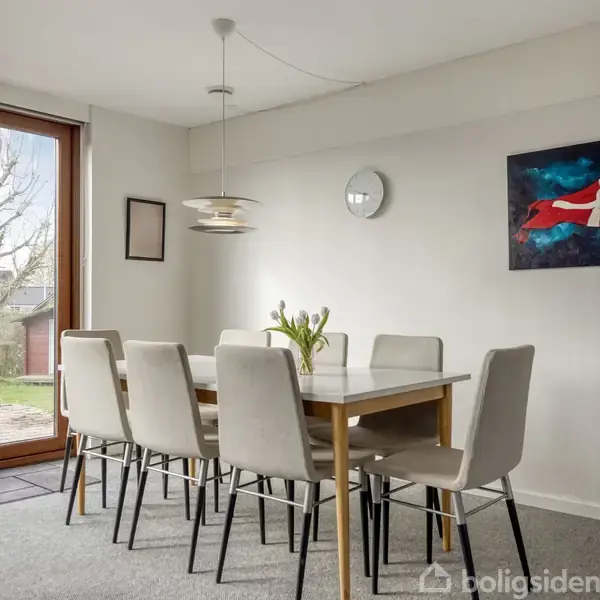 Et spisebord står stille under en moderne lampe med otte polstrede stole rundt om. En vase med blomster pynter bordet, mens en dør åbner ud til en have. Et ur hænger på væggen ved siden af et maleri.