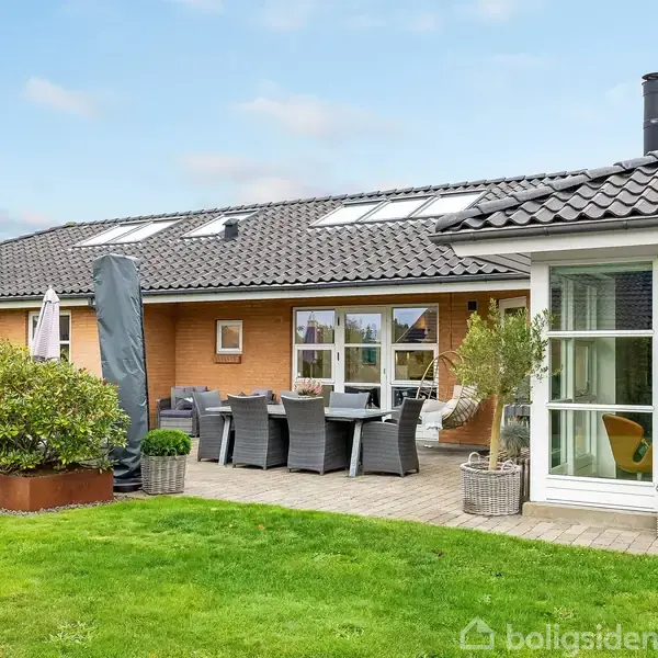 Hus med sort tegltag og gule mursten; havemøbler står på en terrasse med fliser; græsplæne og buske omgiver området.