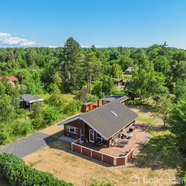 Et rødt enplanshus med gråt tag står omgivet af grønne træer og buske i et landområde, med en terrasse foran og en asfalteret indkørsel til venstre.