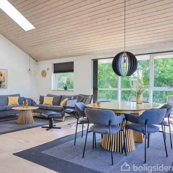 Stue med moderne møbler, herunder blå sofaer og spisebord med stole. Store vinduer i baggrunden giver udsigt til grønne omgivelser. Enkel indretning med lyse trævægge og lofter.