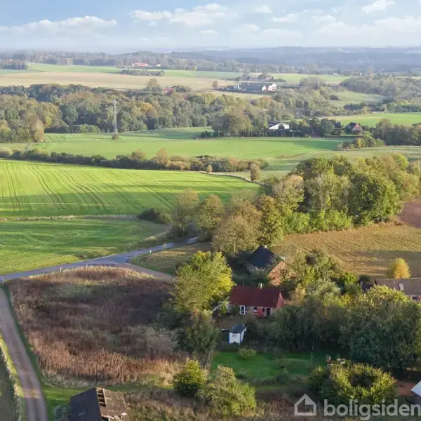 Landsbyhuse omkranset af marker. Husene står stille blandt træer i landlige omgivelser. Marker strækker sig vidt, mens skyer dækker den blå himmel i baggrunden.