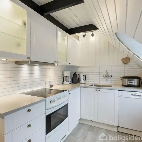 Et hvidt køkken med indbyggede skabe og apparater. En opvaskemaskine, ovn og kogeplader er i brug. Hældende loft og træpaneler giver et hyggeligt, moderne udseende.