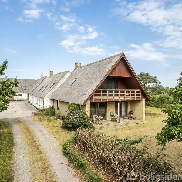 Et stråtækt hus står stille i en landlig omgivelser med grønne træer omkring. En grusvej leder op til huset, og der er blå himmel med spredte skyer.