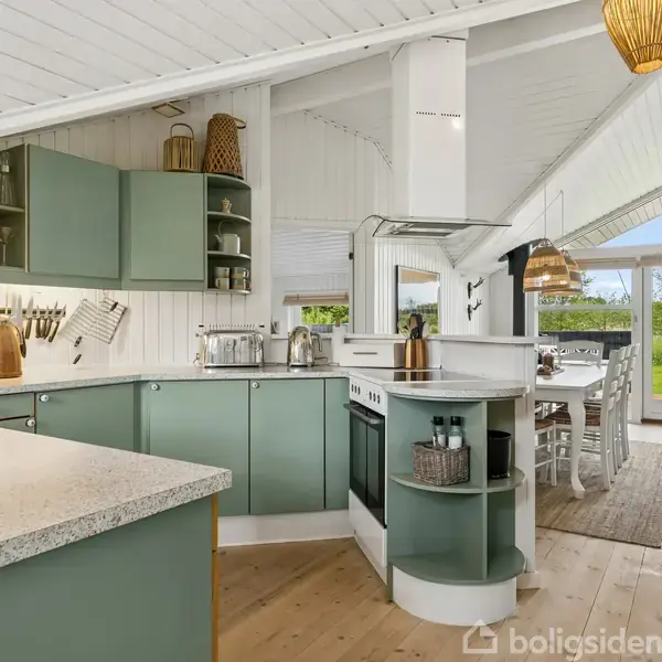 Køkken med grønne skabe og hvide vægge indikerer en moderne stil. Køkkenet fører til et spiseområde med store vinduer, der åbner ud mod en grøn have.