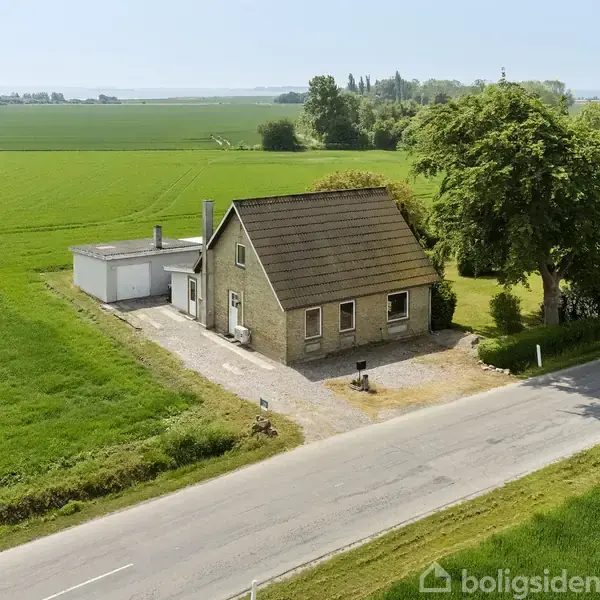 Et lille hus ligger ved en landevej omgivet af grønne marker. En garage er tilknyttet huset. I baggrunden ses træer og en fjern vandudsigt.