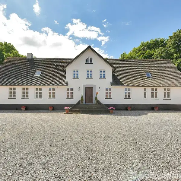 En hvid villa med mørkt tag står på en grusbelagt gårdsplads omgivet af grønne træer. Hoveddøren er i midten med symmetrisk placerede vinduer og blomsterkrukker.