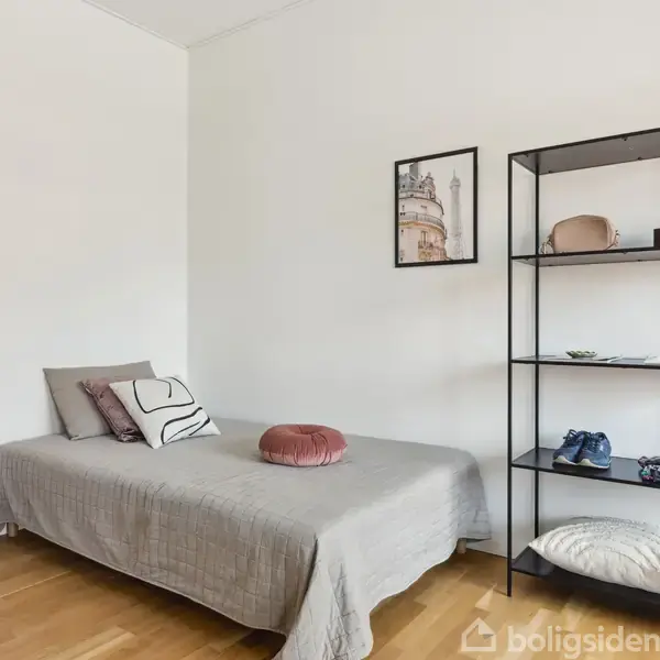 En enkeltseng med gråt sengetæppe står ved en væg, foruden en hylde med bøger og tøj. Et billede hænger over sengen i et lyst værelse med trægulv.