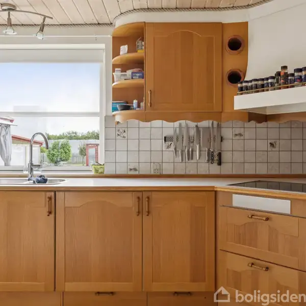 Køkken med lyse træskabe, hvide flisevægge, og stålvaske, med knive ophængt til højre. Udendørs udsyn ses gennem vinduet, omgivet af åbne hylder med krydderier.