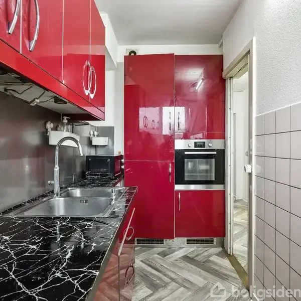 Et smalt køkken med røde skabe og en sort marmorbordplade. Skufferne er åbne, og der hænger køkkenredskaber på væggen. Ovn integreret i skabsmodul. Væggene er delvist flisebelagte.
