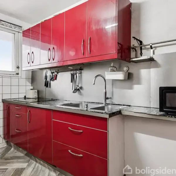 Røde køkkenskabe hænger og står langs væggen, med en håndvask under et vindue. En mikrobølgeovn står til højre på bordpladen. Gulvet er i sildebensmønster.