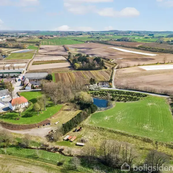 En gård omgivet af grønne marker og en sø ligger i et åbent, landligt landskab med træer og marker i forskellige farver, der strækker sig mod horisonten.