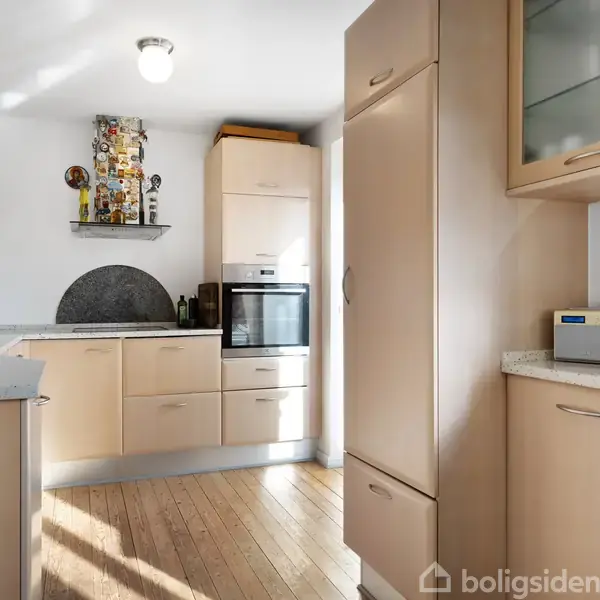 Et moderne køkken med lyse træskabe og indbyggede apparater står i et solbelyst rum med lyse trægulve. Dekorationer og en skål med frugt står på bordpladerne.