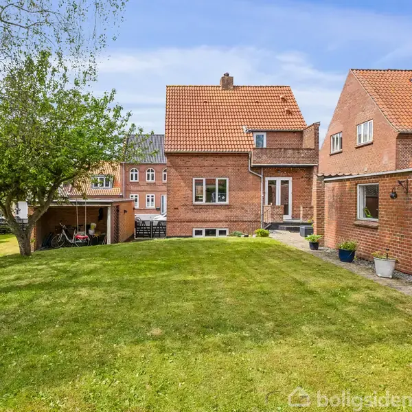 Rød murstenshus står i en have med en grøn græsplæne. En cykelskur ligger til venstre. Huset har flere vinduer og en skorsten. Himlen er blå med få skyer.