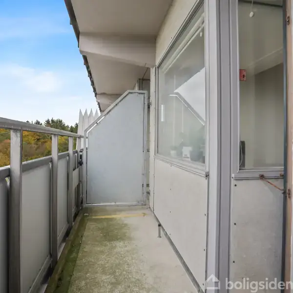 Balkon med metalgelænder strækker sig langs bygningen. Døre og vinduer åbner ud til balkonen. I baggrunden ses trætoppe og himmel under en blå, skyfri dag.