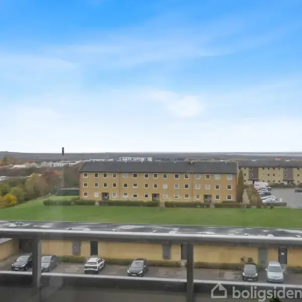 Bygning ses fra en balkon; flere etageboliger står stille i baggrunden. Græsareal og parkeringsplads med biler omgiver bygningerne under en blå himmel.