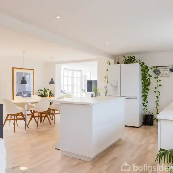 Køkken med hvidt interiør, hvor et køleskab er dækket af grønne planter. I baggrunden ses et spisebord med stole. Rummet er lyst med trægulv og vægdekorationer.