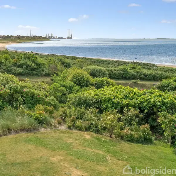 Strand med græs og grønne buske i forgrunden, havet breder sig ud mod horisonten under en klar blå himmel. En kystlinje strækker sig langs venstre side.