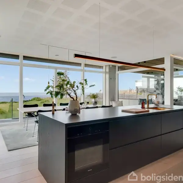 Køkkenø med sort overflade står i forgrunden; bagved ses en spiseplads ved store vinduer, der giver udsigt til en grøn have og havet i baggrunden.