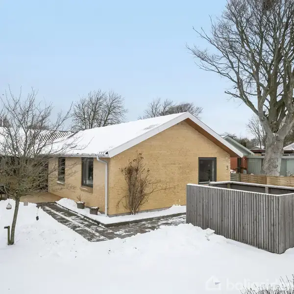 Et gult murstenshus står stille omgivet af snebeklædt jord og spredte træer. Ved siden af huset ses et hegn, og bagved er der flere træer uden blade i vinterlandskabet.