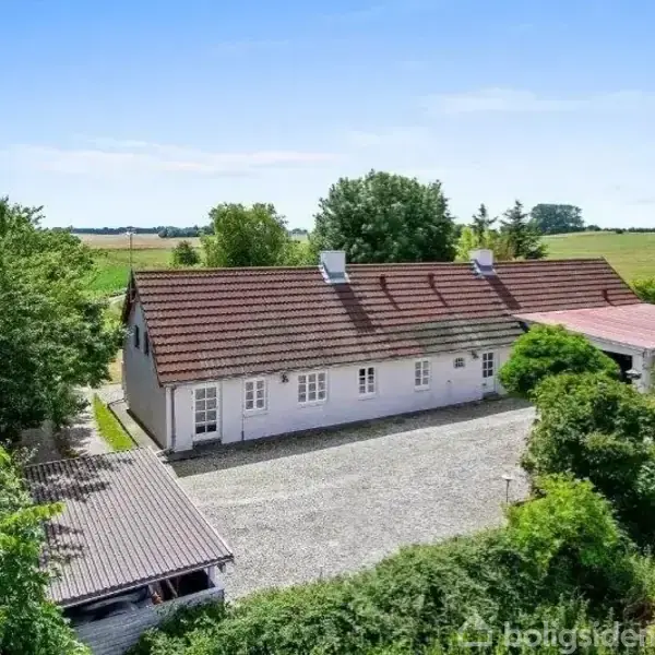 Et hvidt hus med rødt tegltag står stille omgivet af grønne træer og marker. Grussti leder til hovedindgangen, og ladens tag ses til venstre.