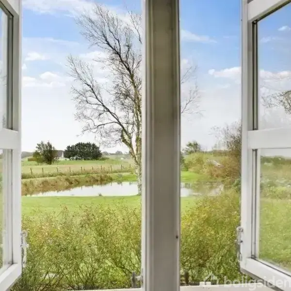 Åbne vinduer, der åbner op til en udsigt over en græsplæne med en lille sø og spredte træer i baggrunden under en blå himmel med lette skyer.