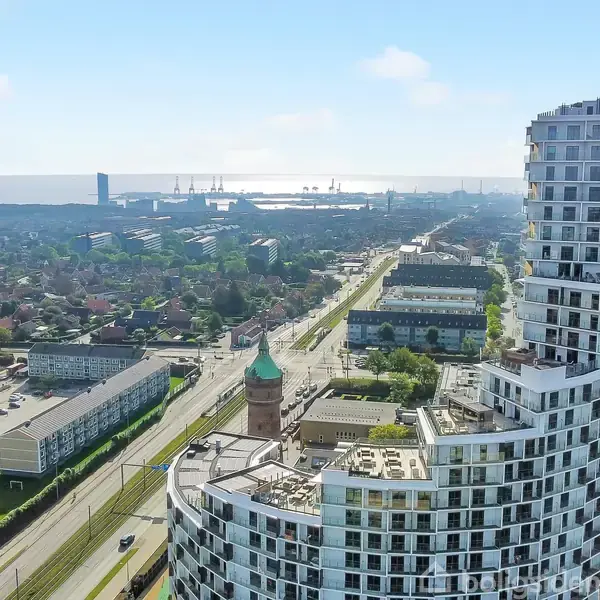 Et højt, moderne lejlighedshus står ved en travl vej med biler. Omkringliggende bygninger og den fjerne havudsigt skaber en storbyatmosfære. Sollys skinner gennem en klar, blå himmel.
