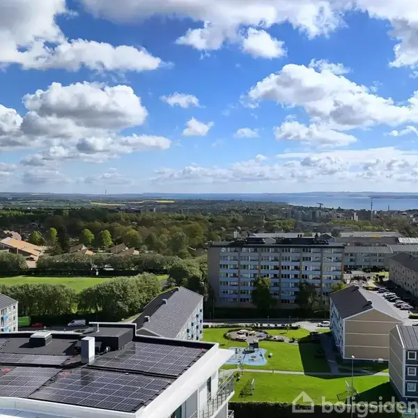 Taget af en bygning med solpaneler under en delvist skyet blå himmel. Bygninger og grønne områder i forgrunden, og havudsigt i baggrunden.
