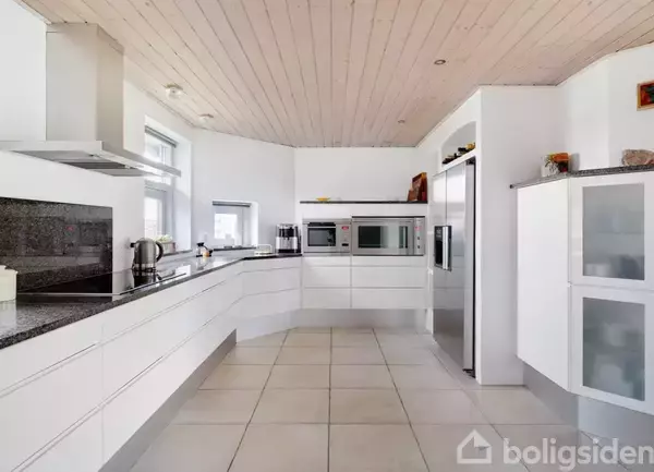 Moderne køkken med hvide skabe, indbyggede ovne og sort granitbordplade. En kedel står på kogepladen. Køleskab og glaslågeskab til højre, lyse gulvfliser og træloft skaber en lys atmosfære.