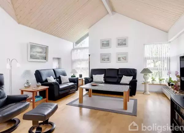 Stue med sorte lædersofaer omkring et træbord, placeret på et trægulv. Væggene har malerier, og store vinduer slipper lys ind. Planter og lamper dekorerer rummet.