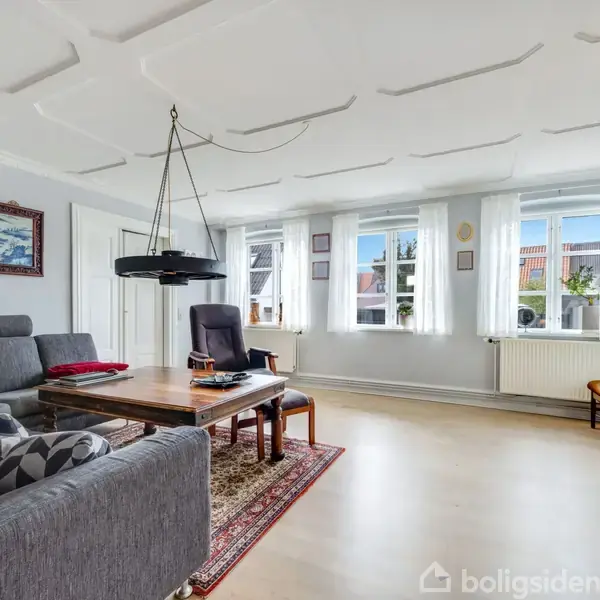 Et moderne opholdsrum indeholder en grå sofa og bord foran et tv. Store vinduer lader naturligt lys ind, mens væggen dekoreres med et maleri.