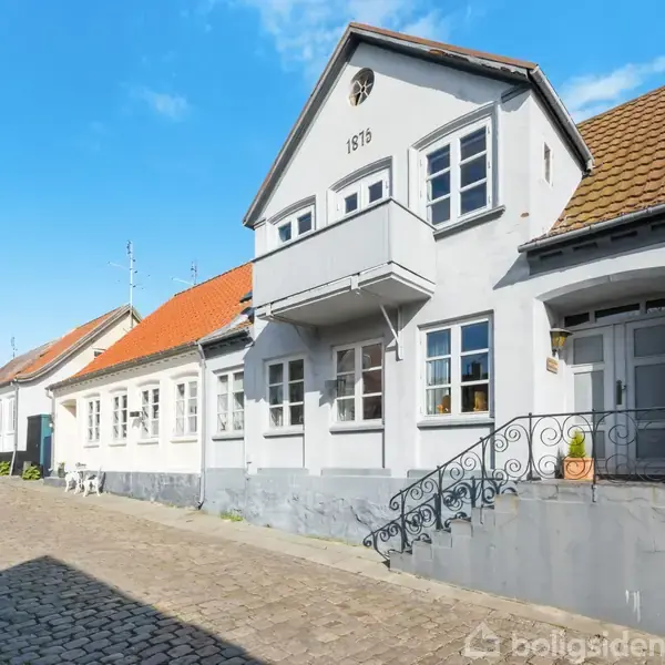 Hus med hvid facade og årstallet "1915", lille balkon, trappe med smedejernsgelænder, beliggende på en brostensgade, omgivet af andre huse i række. Solrigt vejr og blå himmel.