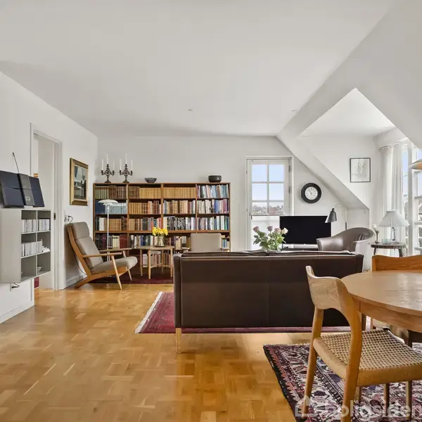 En lys stue med trægulv og spisebord, hvor der er en reol fyldt med bøger. En sofa og lænestol står ved siden af et vindue, omgivet af planter og en lampe.