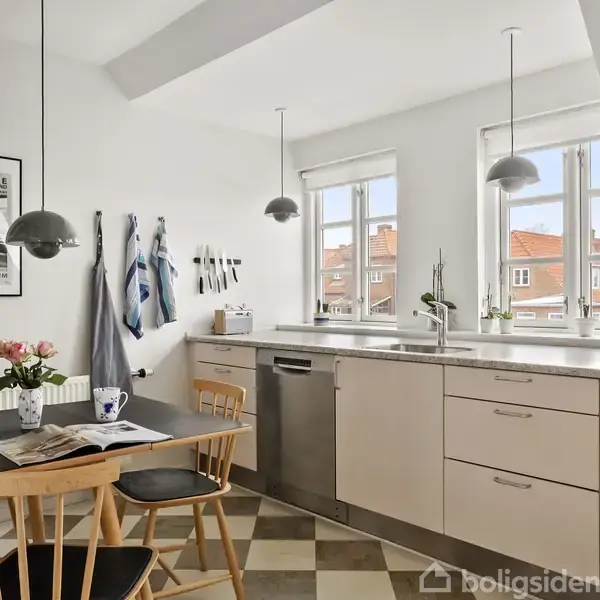 Køkkenbord er fyldt med planter og køkkenredskaber. Stolene står ved et spisebord med en lampe, blomster og en bog. Vinduerne viser røde hustage udenfor. Væg har hængt viskestykker.