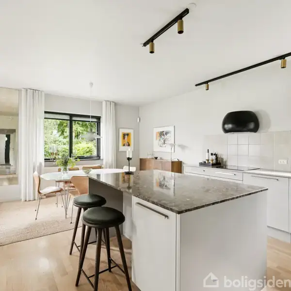 Et moderne køkken med en stor køkkenø der står centralt. Der er hvide skabe, barstole og et spiseområde ved et vindue med udsigt til en grøn have.