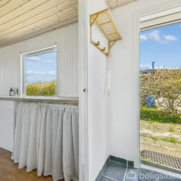 En indgang med et flisebelagt gulv fører til et lyst køkken. Køkkenet har en hvid væg og gardiner under en bordplade. En åben dør viser en have med buske udenfor.