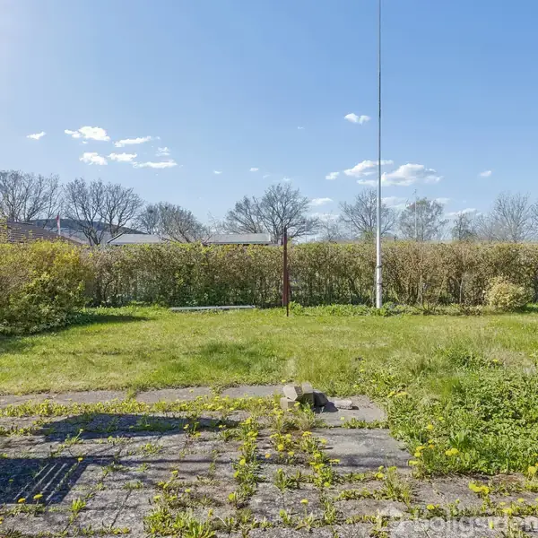 Græsplæne med enkelte fliseområder beliggende i en have. Træer og buske omkranser græsset under en klar blå himmel. En høj, hvid flagstang står centralt.