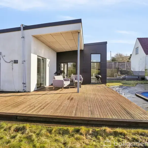En moderne hvid- og sortmalet villa med terrasse har møbler placeret udendørs. Ved siden af terrassen er der en swimmingpool. Nabohuse og en træhegn ses i baggrunden.