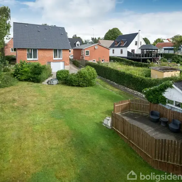 Hus i baggrunden står stille på en velplejet græsplæne omgivet af hække. I forgrunden er en træterrasse med flere sorte stole. Skyer ses på himlen.