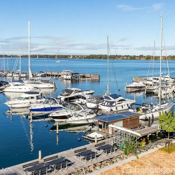 Flere lystbåde ligger fortøjet ved en moderne marina med glitrende blåt vand. I baggrunden ses en bro og kystlinje, mens træer og promenade pryder forgrunden.