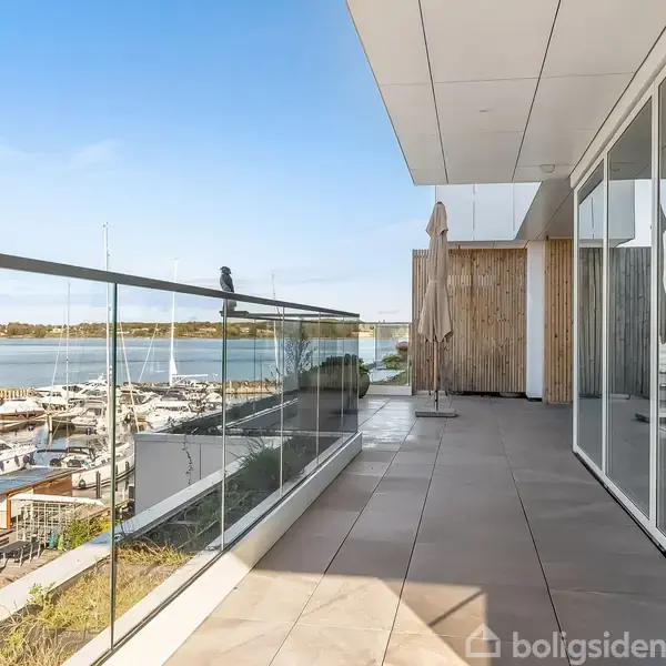 En bred altan med glasrækværk giver udsigt over en marina fyldt med både ved kaj. Sollys skinner på de store gulvfliser, og terrassen har moderne glasdøre.