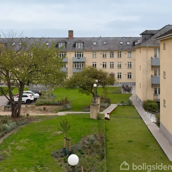 Beboelsesbygninger omringer en græsplæne med træer og belysning. Parkerede biler ses på grusområder, og der er cykler i det fjerne. Himmel bagved er overskyet.