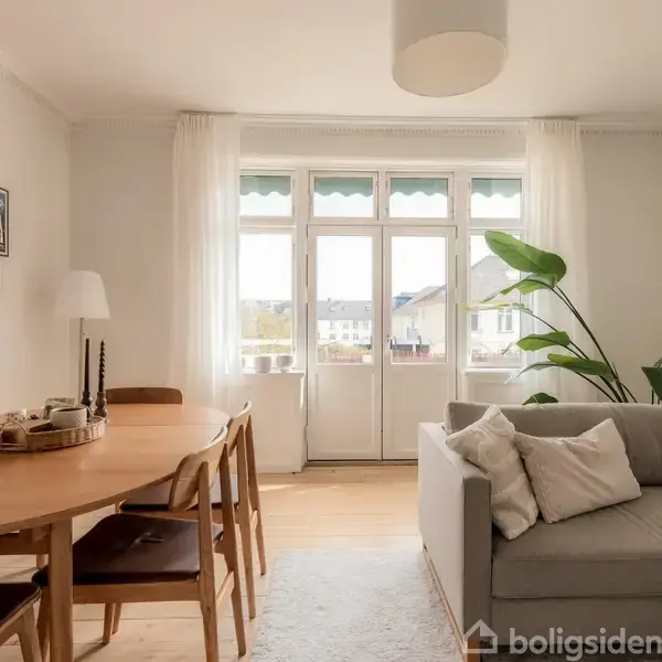 En lys stue med en grå sofa til højre, et rundt spisebord med træstole til venstre, store vinduer med hvidt gardin mod bagvæggen, og vægdekorationer på venstre væg.