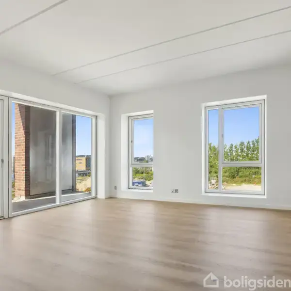 Et lyst, tomt værelse med stort trægulv og tre store vinduer, der giver udsigt til et moderne byggeri og grønne områder udenfor.