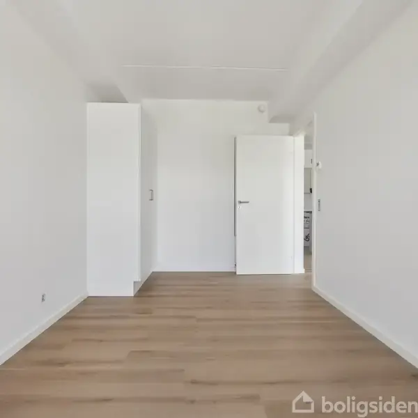 Et tomt rum med en hvid dør åbner ind til et andet rum. Trægulvet strækker sig over det lyse rum, og hvide vægge omgiver pladsen. Ingen tekst til stede.